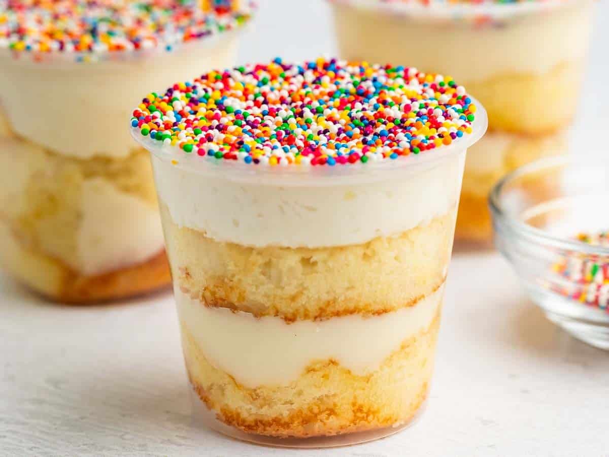 A clear plastic cup filled with layers of vanilla cake and creamy frosting, topped with colorful rainbow sprinkles. Two similar cups and a glass bowl of sprinkles are in the background.