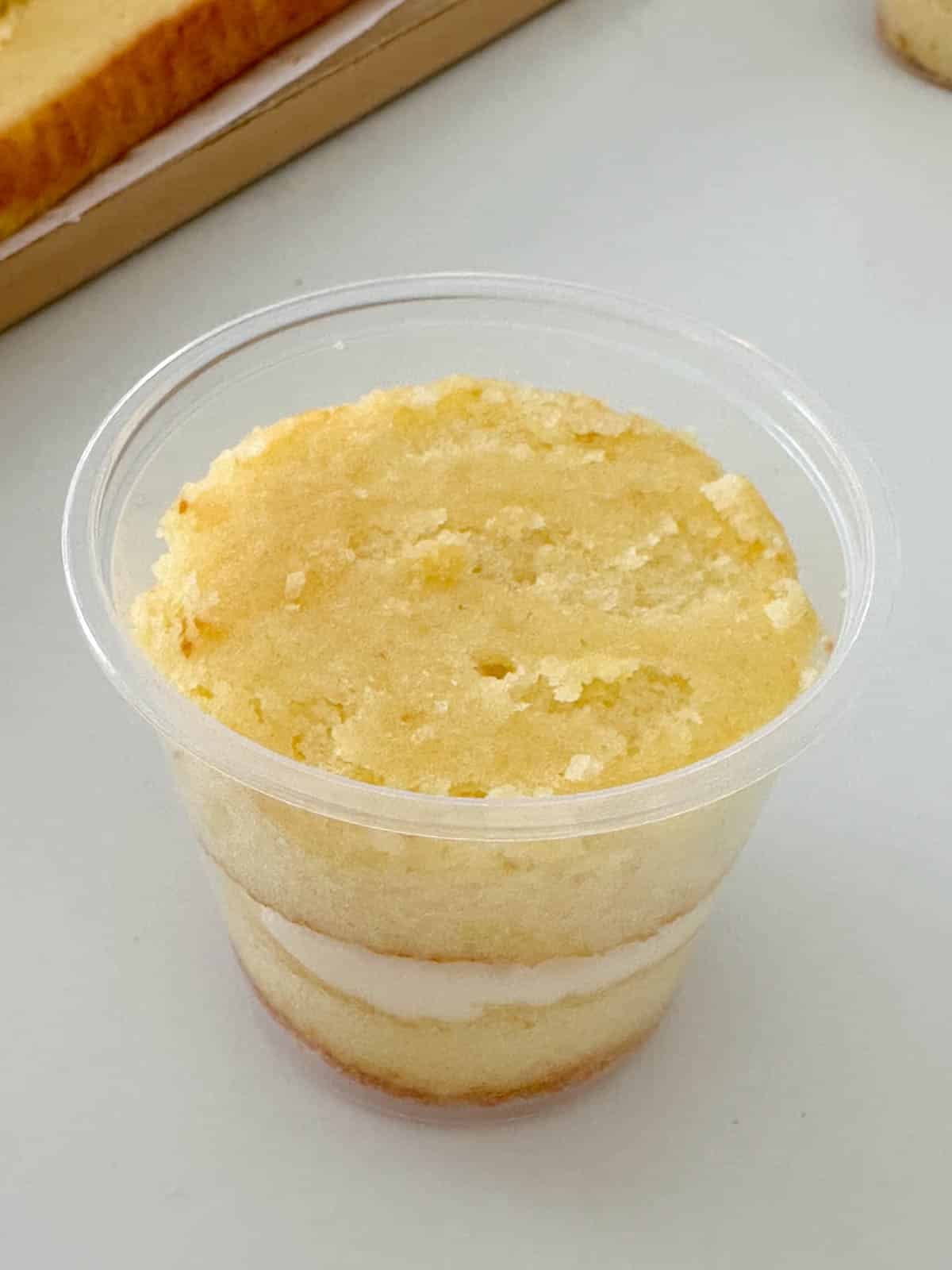 A clear plastic cup containing a single-serving layered vanilla cake with a layer of white frosting in the middle, placed on a light-colored surface.