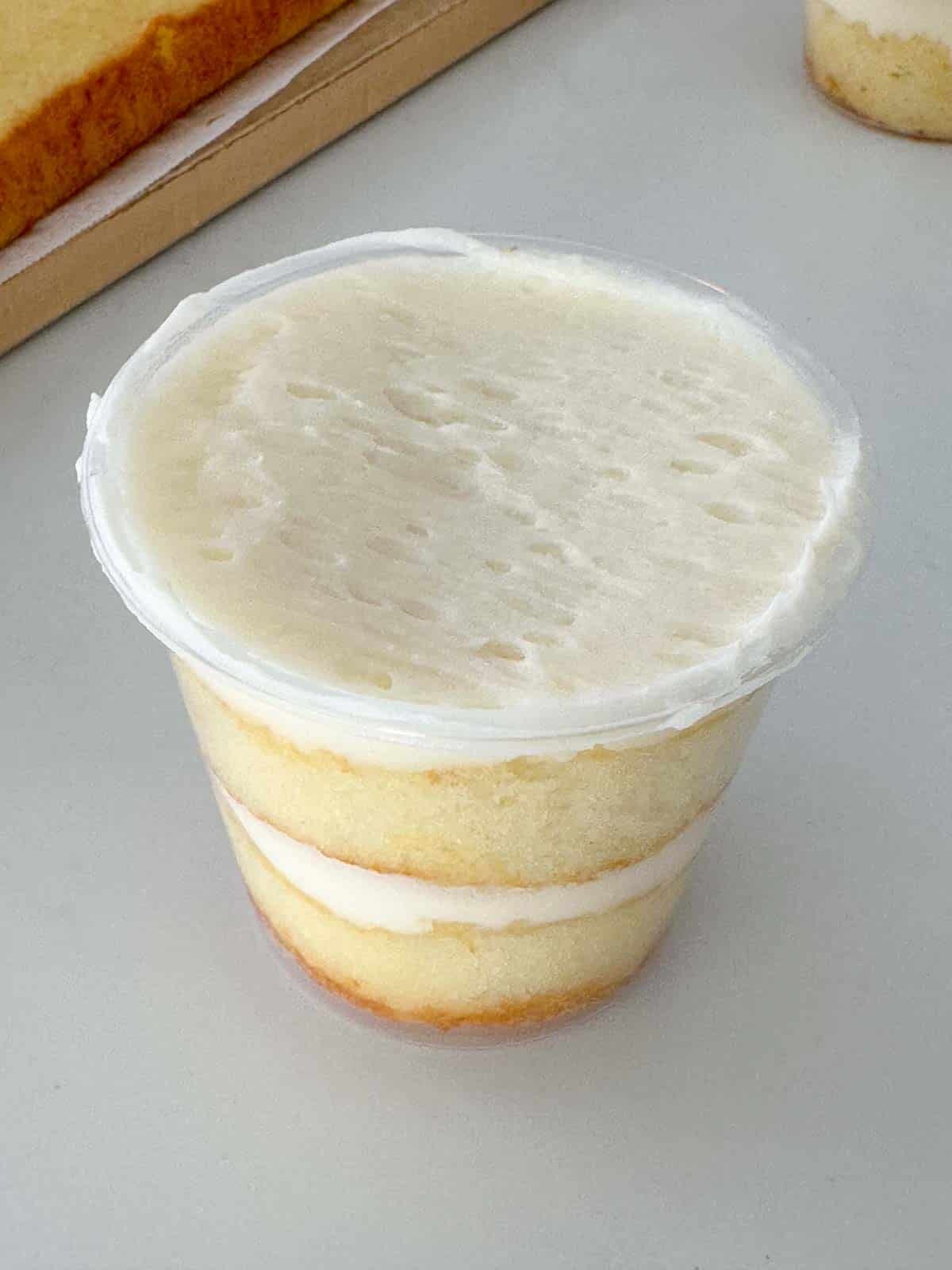 A clear plastic cup filled with layers of vanilla cake and white frosting, with a smooth layer of frosting on top. The cup is placed on a light-colored surface.