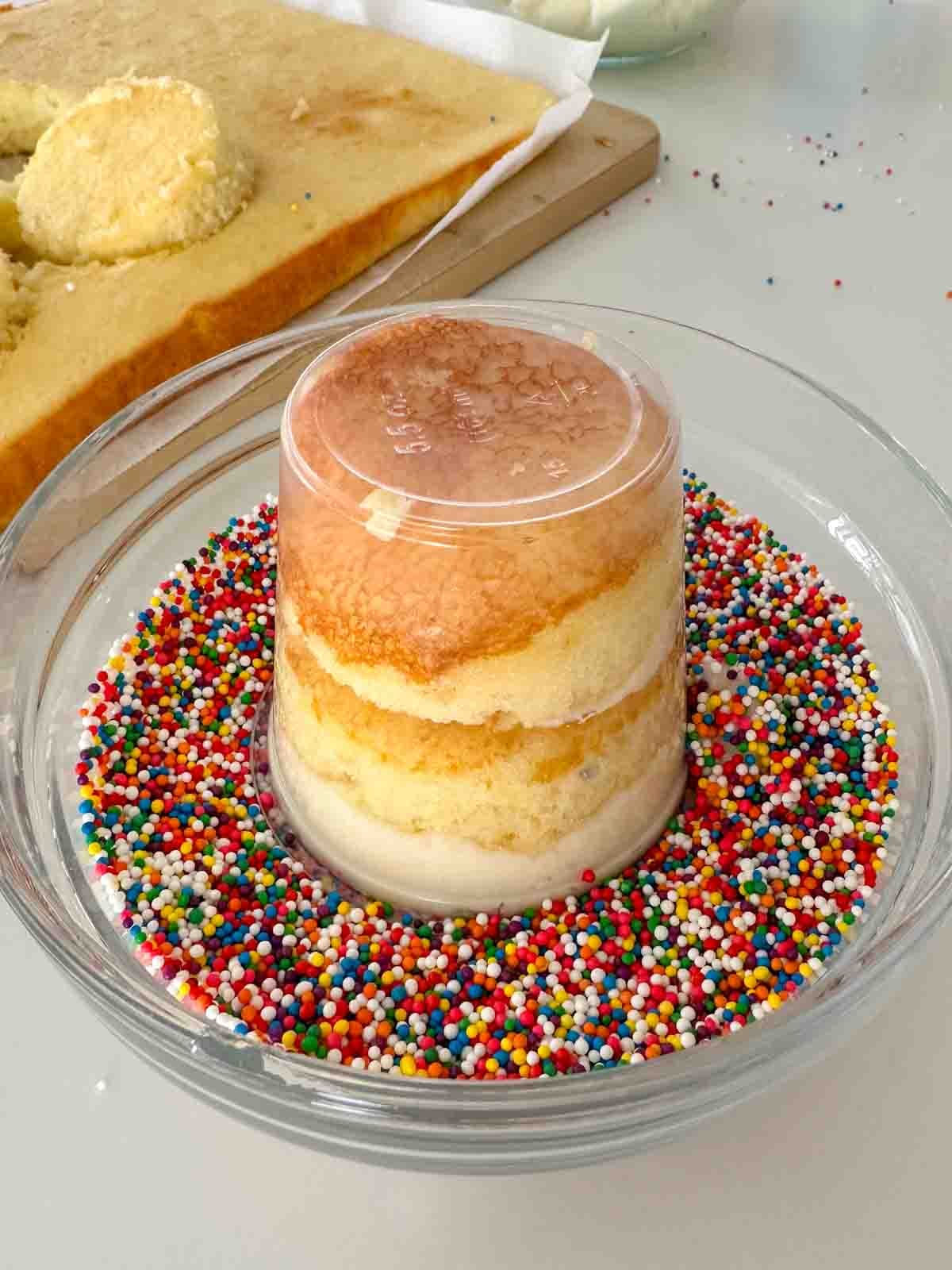 A small, round, layered cake sits upside down in a bowl filled with colorful round sprinkles, with a sheet cake in the background.