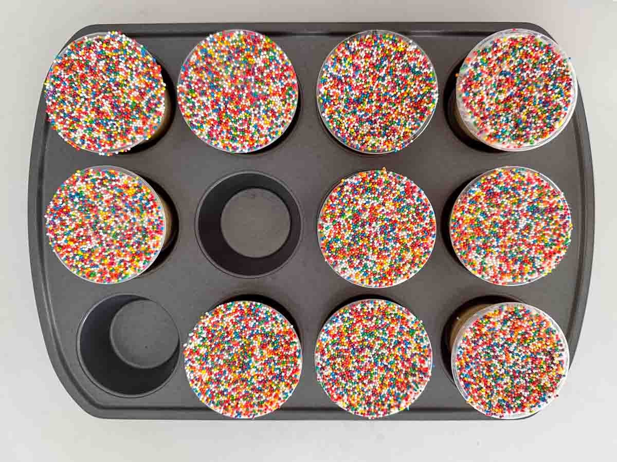 A muffin tin holds ten cupcakes topped with colorful round sprinkles, with two empty slots visible in the pan.