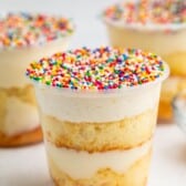 A clear plastic cup filled with layered vanilla cake and cream, topped with white frosting and colorful rainbow sprinkles. Two similar dessert cups appear blurred in the background.