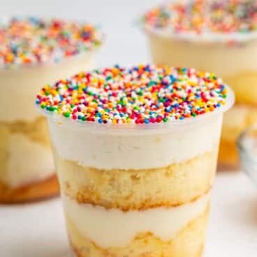 A clear plastic cup filled with layered vanilla cake and cream, topped with white frosting and colorful rainbow sprinkles. Two similar dessert cups appear blurred in the background.