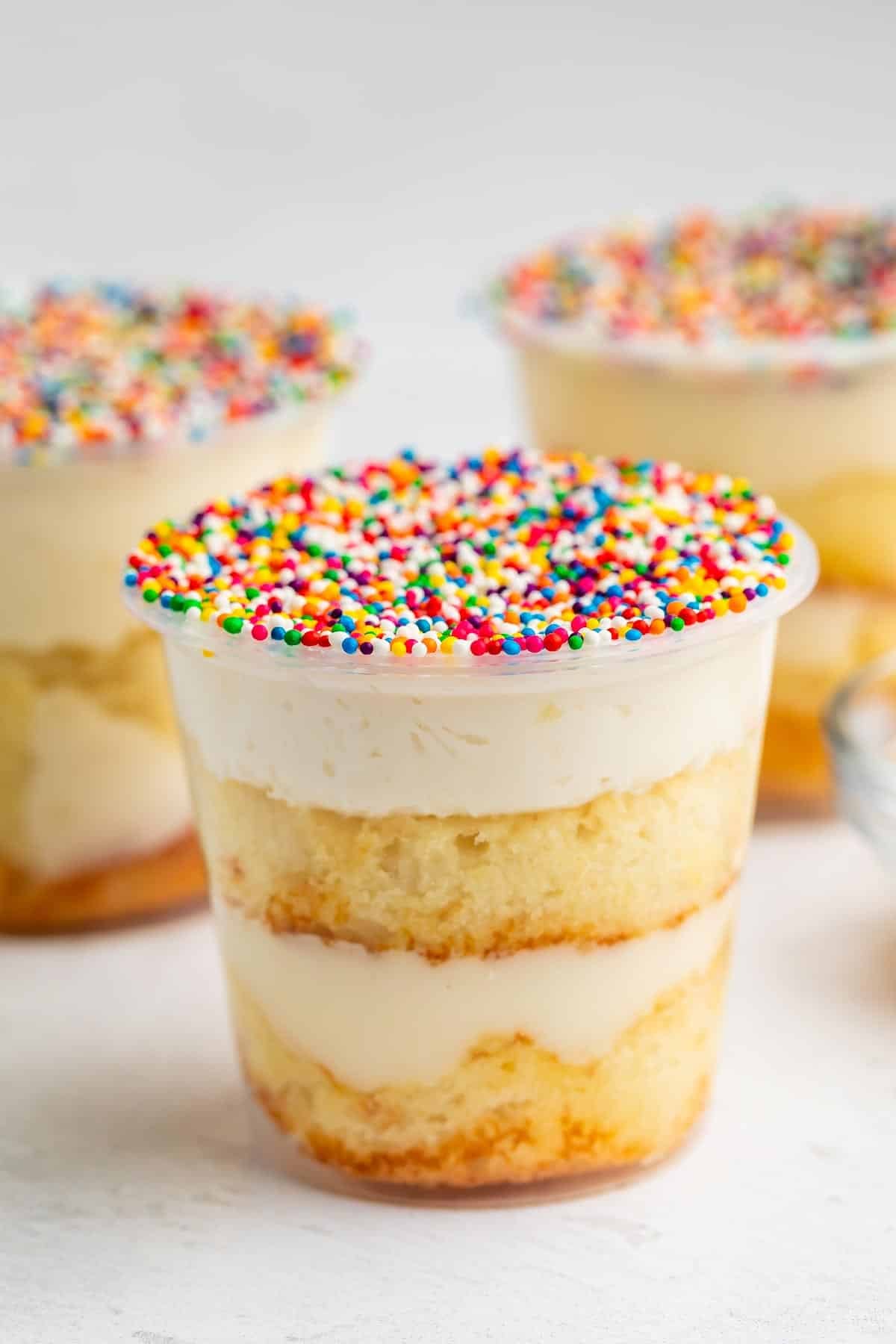 A clear plastic cup filled with layered vanilla cake and cream, topped with white frosting and colorful rainbow sprinkles. Two similar dessert cups appear blurred in the background.