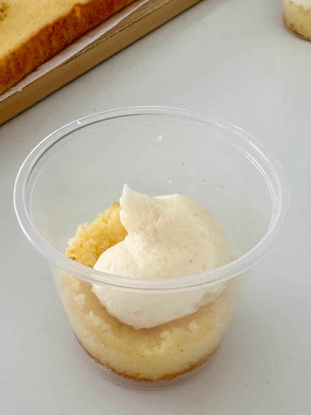 A clear plastic cup containing a layer of yellow cake topped with a dollop of white frosting, sitting on a white surface. Another piece of cake is partially visible in the background.