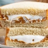 A close-up of a sandwich cut in half, showing layers of creamy peanut butter and fluffy white marshmallow spread between two slices of white bread. The sandwich sits on a plate.