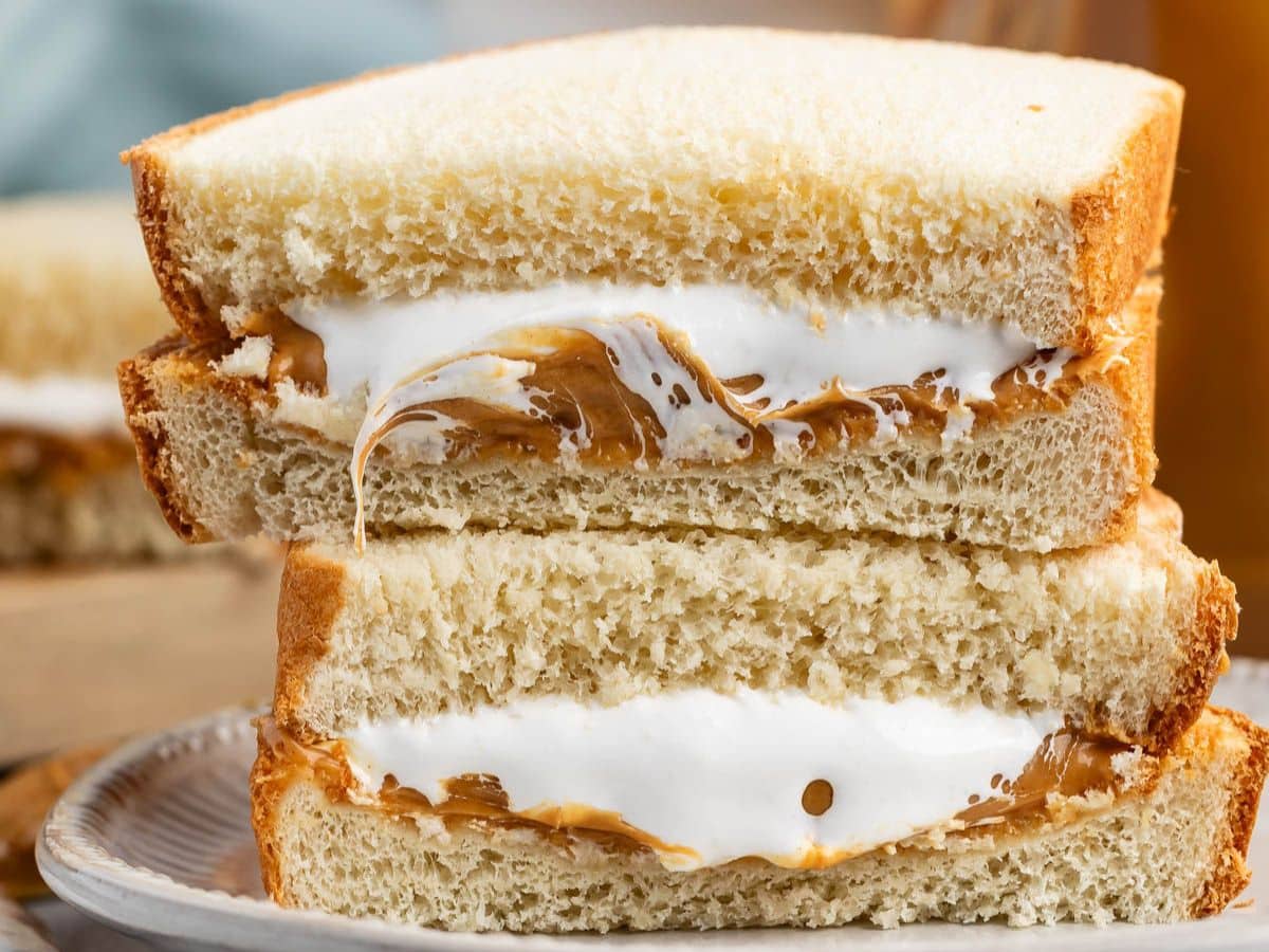 A close-up of a sandwich cut in half, showing layers of creamy peanut butter and fluffy white marshmallow spread between two slices of white bread. The sandwich sits on a plate.