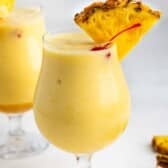 A creamy yellow piña colada served in a curved glass, garnished with a pineapple wedge and a red cherry. Another similar drink is blurred in the background on a light surface.