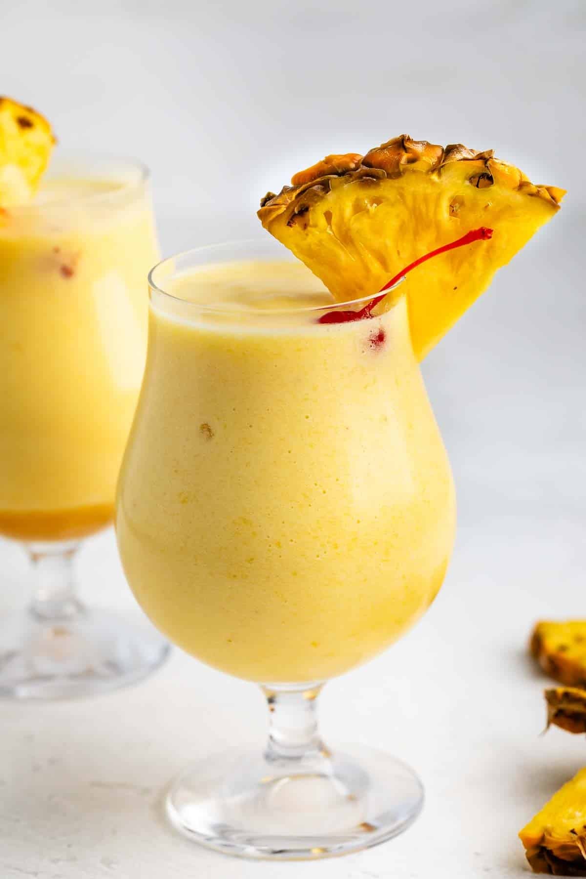A creamy yellow piña colada served in a curved glass, garnished with a pineapple wedge and a red cherry. Another similar drink is blurred in the background on a light surface.