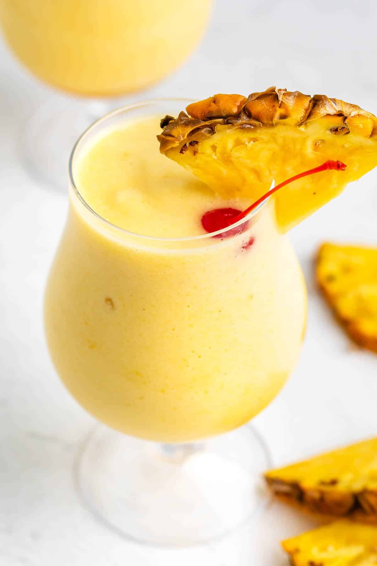 A creamy yellow piña colada in a curved glass, garnished with a pineapple slice and a red cherry. Pineapple wedges are scattered on the white surface in the background.