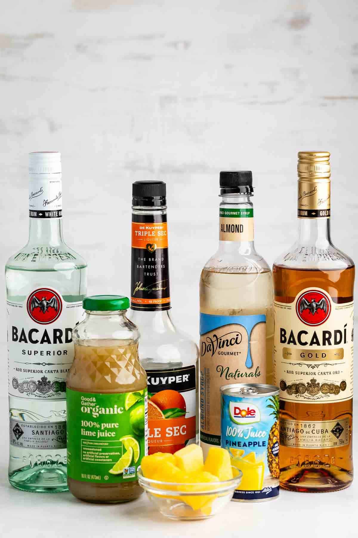 Five bottles of drink ingredients, a jar of lime juice, a can of pineapple juice, and a bowl of pineapple chunks are arranged on a white surface with a light gray background.