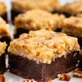 A close-up of a chocolate brownie bar topped with a thick layer of coconut and pecan frosting, surrounded by scattered pecan pieces on a white surface.