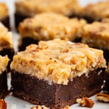 A close-up of a chocolate brownie bar topped with a thick layer of coconut and pecan frosting, surrounded by scattered pecan pieces on a white surface.