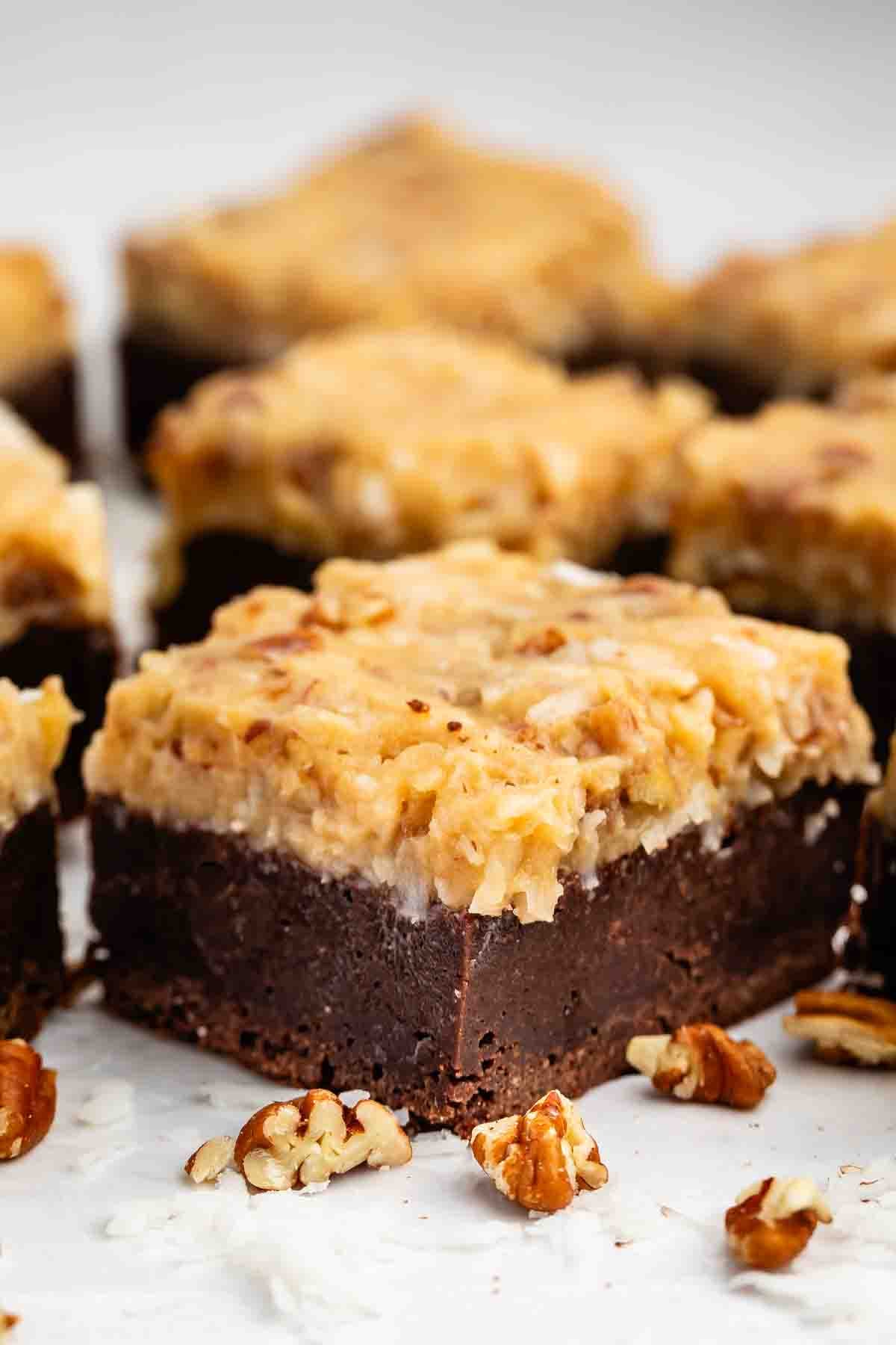 A close-up of a chocolate brownie bar topped with a thick layer of coconut and pecan frosting, surrounded by scattered pecan pieces on a white surface.