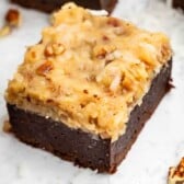 A square piece of German chocolate brownie topped with a coconut and pecan frosting, surrounded by small bits of nuts on a white surface.