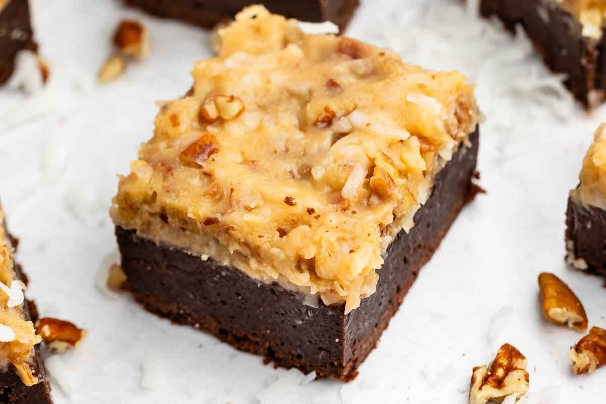 Un morceau carré de brownie au chocolat allemand garni de noix de coco et de pacanes, entouré de petits morceaux de noix sur une surface blanche.