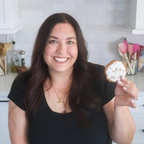 About: A woman with long dark hair, wearing a black shirt, smiles and holds up a cookie with white icing in a bright kitchen with white cabinets and utensils in the background.