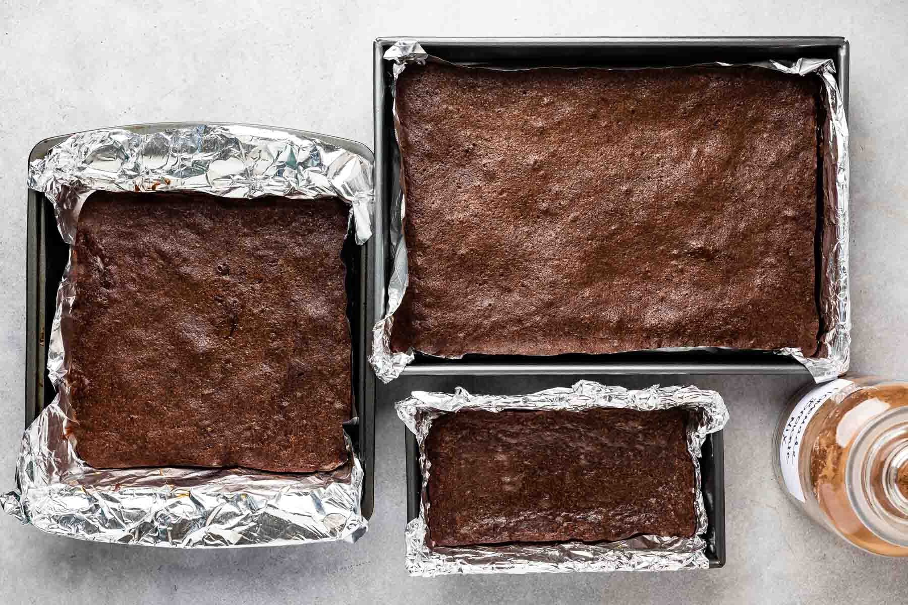 Three baking pans lined with aluminum foil, each filled with uncut brownies made from homemade brownie mix, sit on a light surface. A bottle with a brown label and white cap is partially visible in the top right corner.