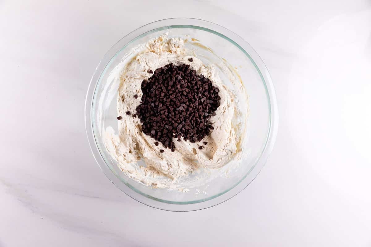A glass bowl filled with creamy cookie dough topped with a pile of chocolate chips, placed on a white marble surface.