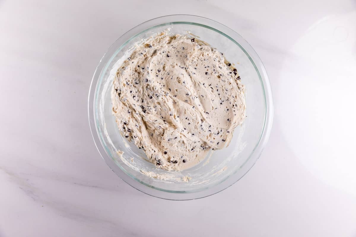 A glass bowl filled with chocolate chip cookie dough sits on a white marble surface, viewed from above. The dough is creamy with visible chocolate chips mixed throughout.