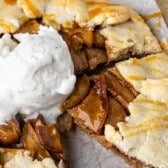 A rustic apple galette with golden crust, filled with cinnamon-spiced apple slices, topped with a scoop of vanilla ice cream and drizzled with caramel sauce. A slice has been cut and pulled slightly away.