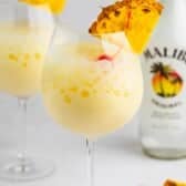 A glass of creamy yellow piña colada garnished with a pineapple slice and cherry. Another glass and a Malibu rum bottle are blurred in the background, with pineapple pieces on the table.