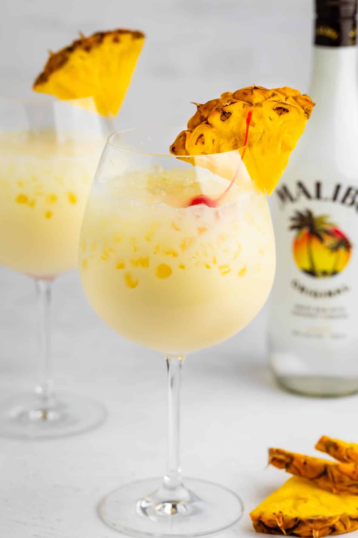 A glass of creamy yellow piña colada garnished with a pineapple slice and cherry. Another glass and a Malibu rum bottle are blurred in the background, with pineapple pieces on the table.