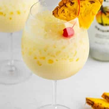 A glass of creamy yellow piña colada with ice, garnished with a pineapple slice and a red cherry, sits on a white surface. A bottle and another glass are blurred in the background. Pineapple pieces are nearby.