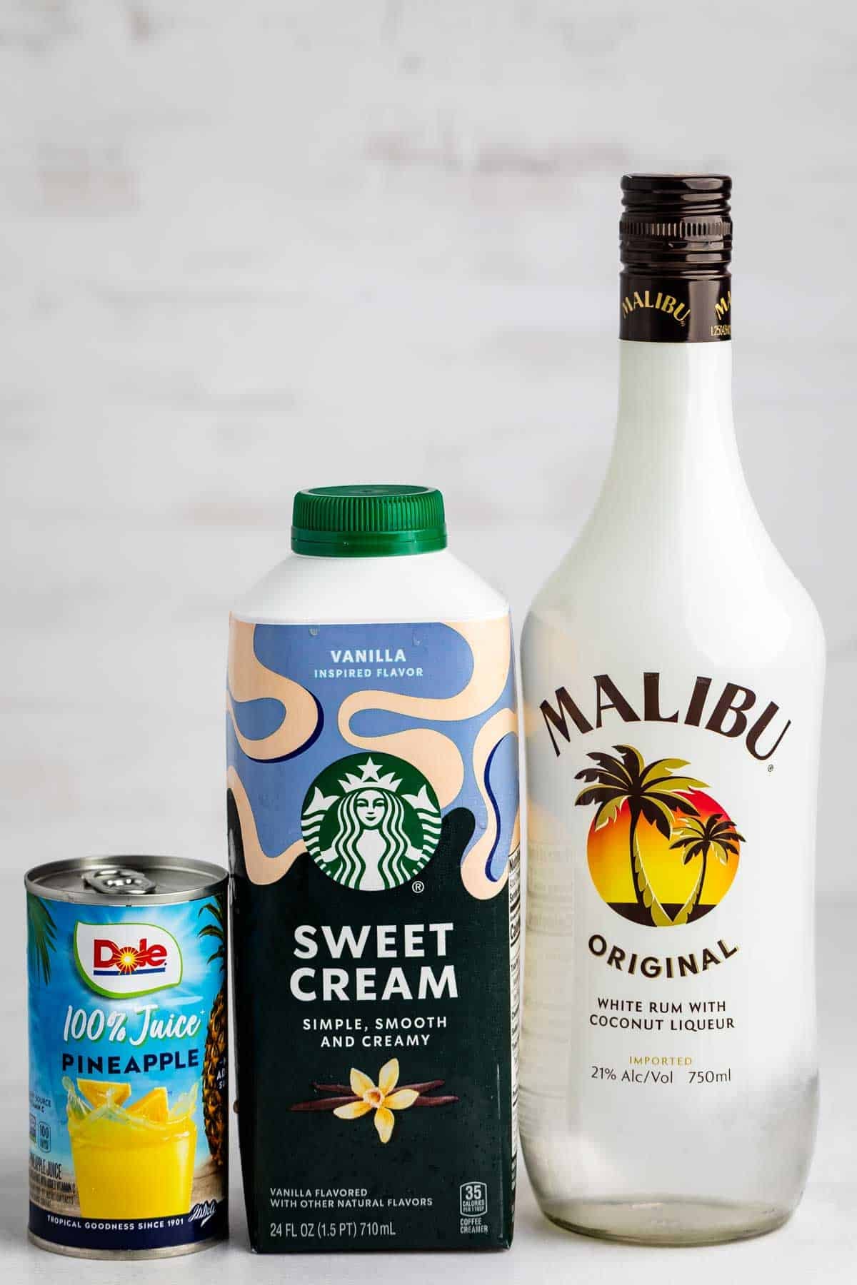 A can of Dole 100% pineapple juice, a carton of Starbucks sweet cream, and a bottle of Malibu Original coconut rum are arranged side by side on a light surface.