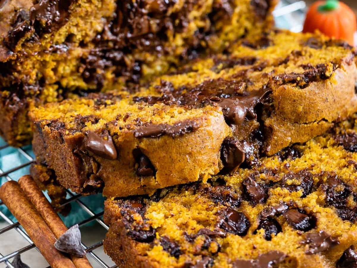 Close-up of sliced pumpkin bread with melted chocolate chips on a cooling rack, with cinnamon sticks and a small decorative pumpkin in the background.