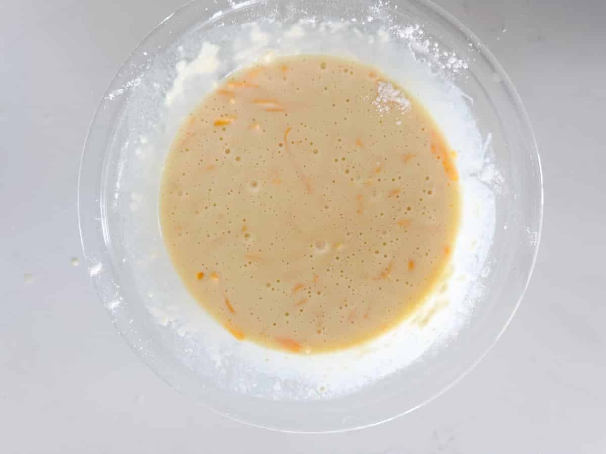 A clear glass bowl filled with pale yellow batter, with small orange shreds mixed in, sits on a white surface. Some flour is visible on the rim of the bowl.