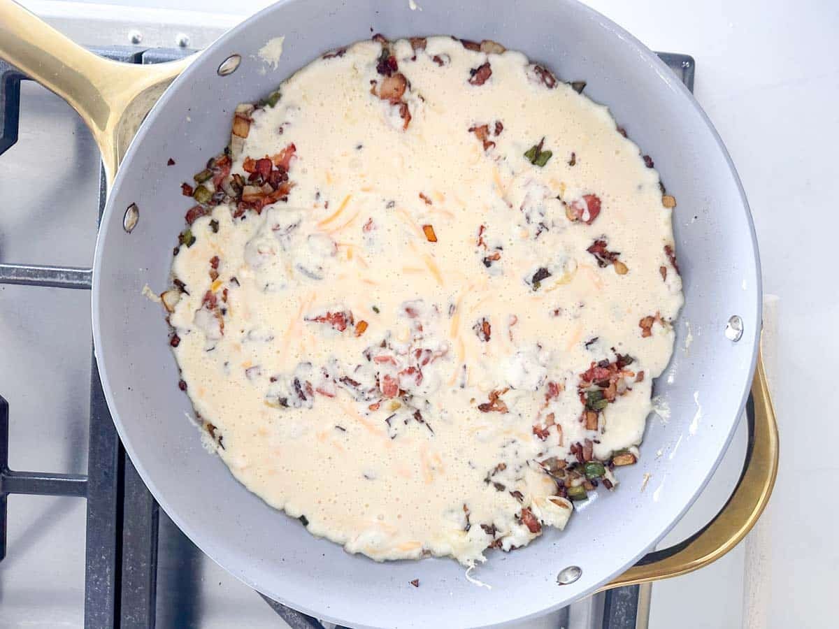 A frying pan on a stove contains a mixture of eggs, chopped vegetables, and cheese, partially cooked and starting to set around the edges. The skillet has a gold-colored handle.