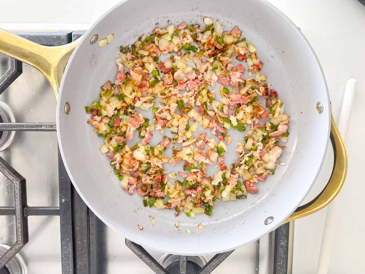 A frying pan on a stovetop with chopped onions, green peppers, and bacon pieces being sautéed together. The ingredients are lightly browned and mixed.