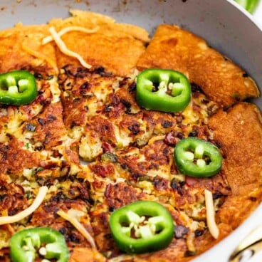 A skillet filled with a golden-brown baked dish topped with melted cheese, chopped ingredients, and sliced fresh jalapeños. A bowl of shredded cheese and a whole jalapeño are in the background.
