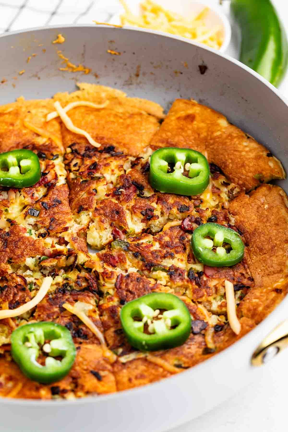 A skillet filled with a golden-brown baked dish topped with melted cheese, chopped ingredients, and sliced fresh jalapeños. A bowl of shredded cheese and a whole jalapeño are in the background.