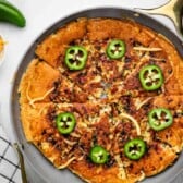 A skillet pizza is topped with melted cheese, browned bits, and sliced jalapeños. A bowl of shredded cheddar, a jalapeño pepper, and a checkered towel are nearby on a white surface.