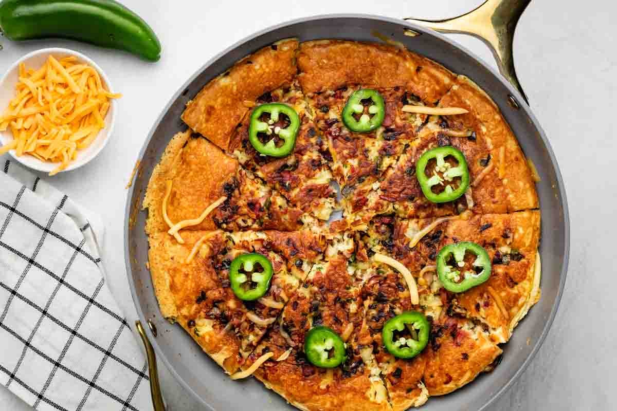 A skillet pizza is topped with melted cheese, browned bits, and sliced jalapeños. A bowl of shredded cheddar, a jalapeño pepper, and a checkered towel are nearby on a white surface.