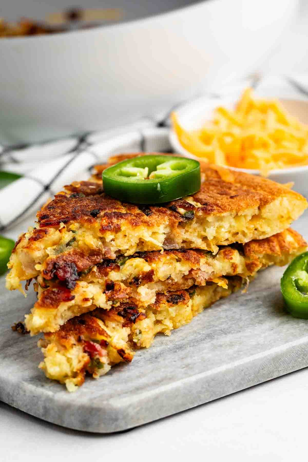 Three stacked slices of golden, crispy cornbread with visible bits of bacon and jalapeño, garnished with a fresh jalapeño slice, served on a gray board with shredded cheese in the background.