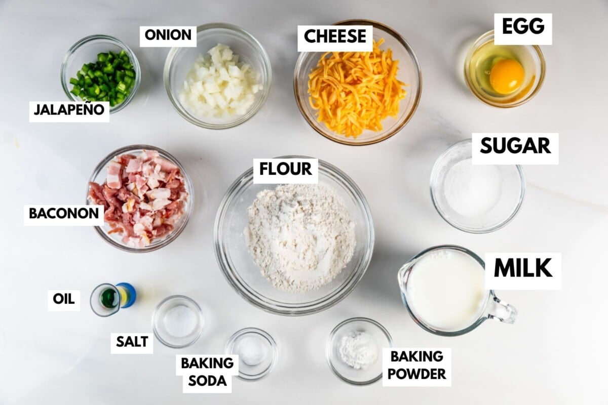 Top-down view of labeled ingredients in separate bowls on a white surface, including flour, shredded cheese, egg, chopped onion, jalapeno, bacon, sugar, milk, oil, salt, baking soda, and baking powder.