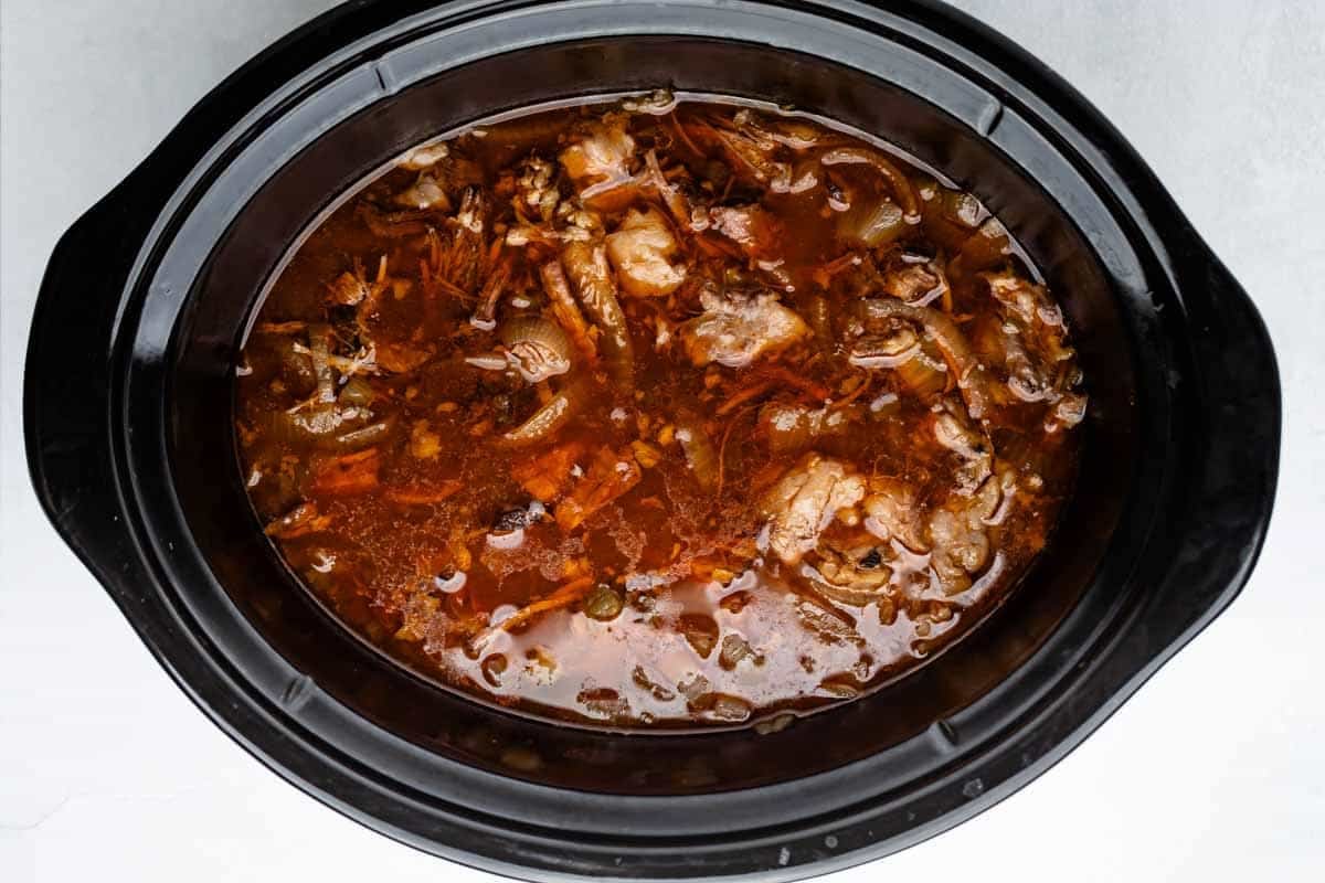 Tacos au bœuf râpé à la mijoteuse 16 Une mijoteuse noire remplie de bouillon brun foncé, de morceaux de viande et de légumes croustillants, placée sur une surface blanche.