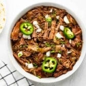 A bowl of shredded beef topped with chopped onions, sliced jalapeños, and fresh herbs, next to a plate with tortillas, on a white surface with a checkered cloth.