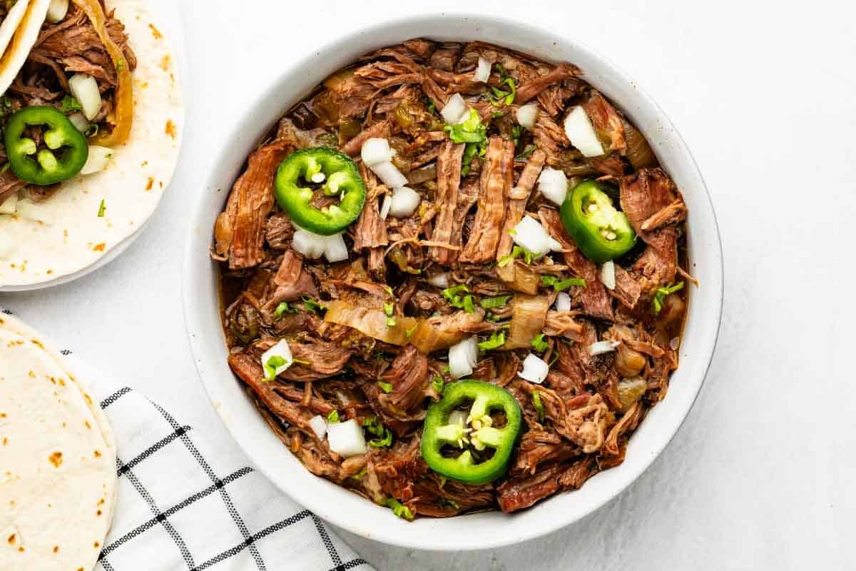 Tacos au bœuf râpé à la mijoteuse 7 Un bol de bœuf râpé garni d'oignons hachés, de tranches de jalapenos et d'herbes fraîches, à côté d'une assiette de tortilla, sur une surface en tissu carré blanc.