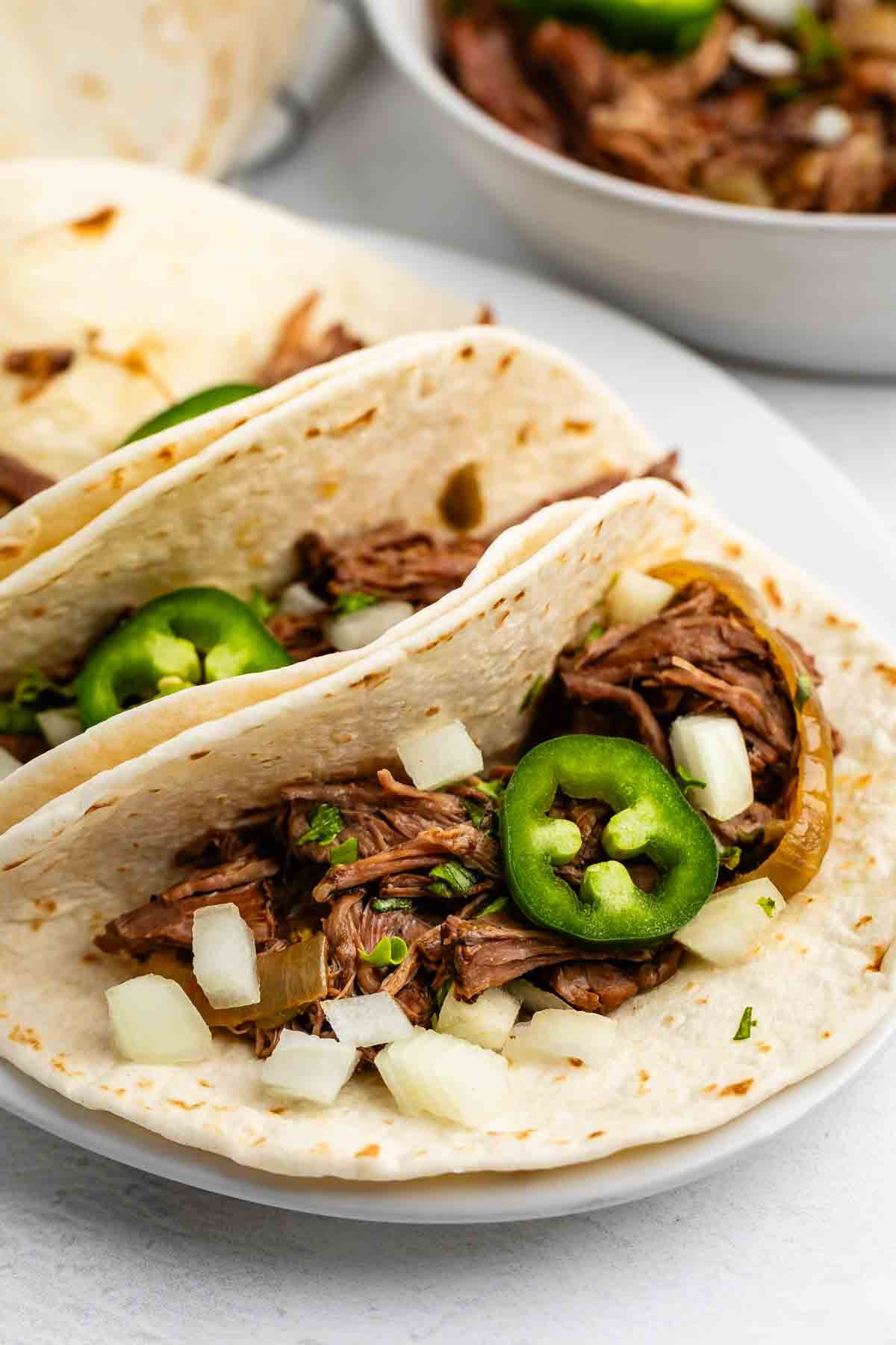Tacos au bœuf râpé à la mijoteuse 12 Deux tacos à la farine molle remplis de bœuf râpé, d'oignons hachés, de coriandre hachée et de jalapenos tranchés, servis sur une assiette blanche. Un bol de bœuf râpé supplémentaire apparaît en arrière-plan.