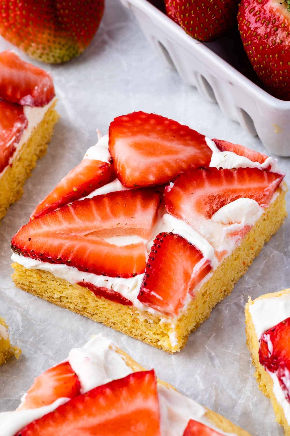 A square piece of dessert with a golden cookie crust, topped with a layer of white cream and sliced fresh strawberries. Other pieces and a container of whole strawberries are visible nearby.