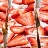 Rectangular dessert bars topped with a layer of white frosting and fresh sliced strawberries, arranged closely together on a tray.