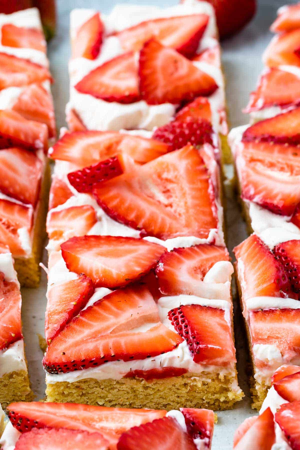 Rectangular dessert bars topped with a layer of white frosting and fresh sliced strawberries, arranged closely together on a tray.