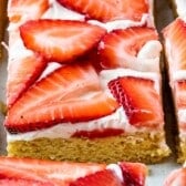 Close-up of dessert bars topped with a layer of white frosting and fresh strawberry slices on a golden-brown base, cut into squares and arranged closely together.