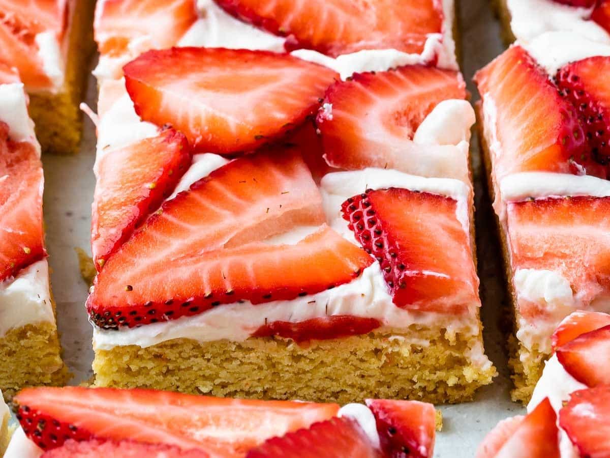 Close-up of dessert bars topped with a layer of white frosting and fresh strawberry slices on a golden-brown base, cut into squares and arranged closely together.
