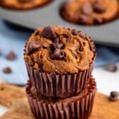Two chocolate chip muffins stacked on a wooden board, with more muffins in a baking tray in the background and scattered chocolate chips around.