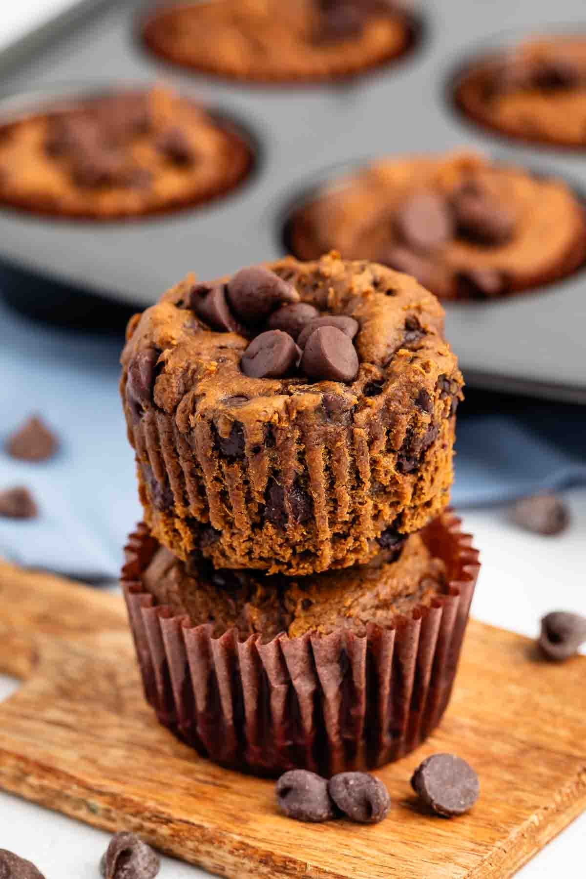 Two chocolate chip muffins are stacked on a wooden board with extra chocolate chips on top. More muffins and scattered chocolate chips are visible in the background.